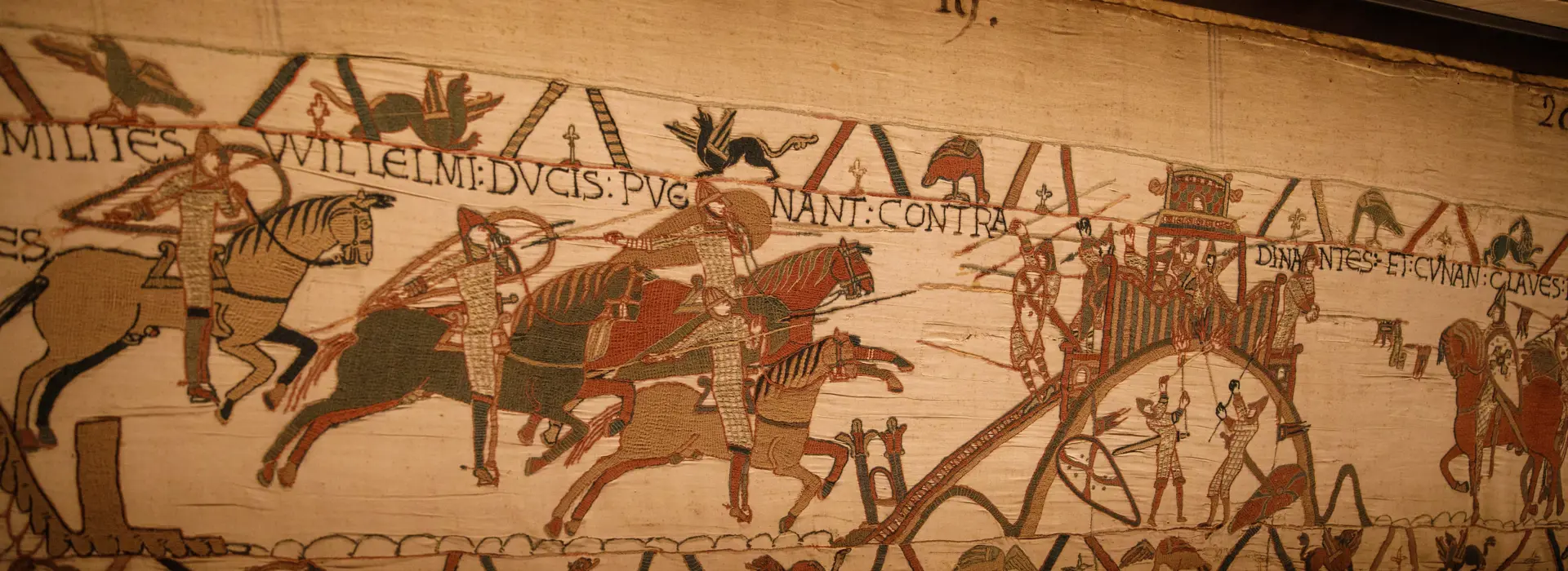 Particolare dell’Arazzo di Bayeux con una scena di battaglia: cavalieri normanni armati di lance e scudi avanzano a cavallo, mentre altri combattenti e un carro trainato da cavalli compaiono sulla destra. Sopra le figure corre un’iscrizione latina ricamata.